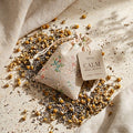 A small beige linen drawstring sachet with floral embroidery sits on a linen surface, surrounded by loose dried chamomile and lavender buds. A tag on the sachet reads 'devas CALM HERBAL INFUSION'.
